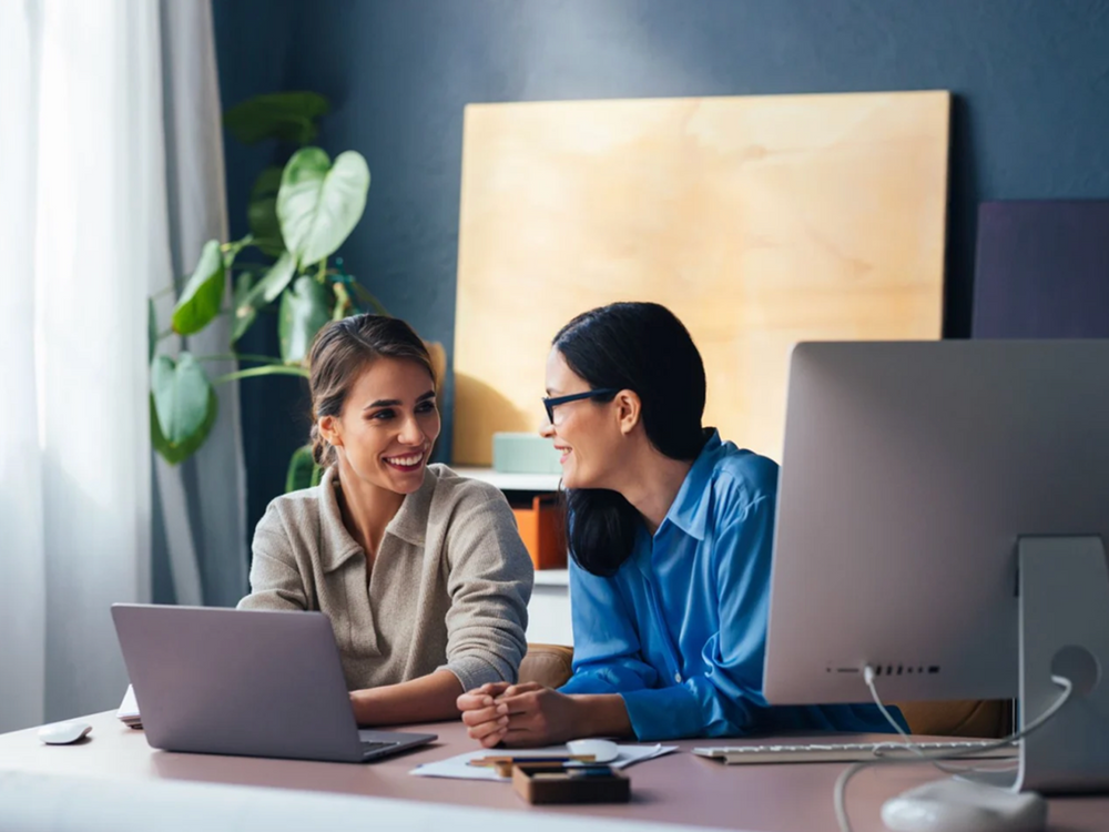 Two women sitting at a desk with a laptop and computer monitor, smiling and engaged in conversation.